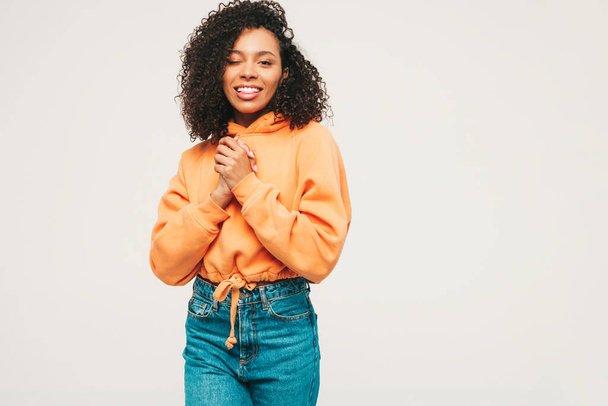 Beautiful black woman with afro curls hairstyle.Smiling model in orange hoodie and trendy jeans clothes. Sexy carefree female posing on white background in studio. Tanned and cheerful - 写真・画像