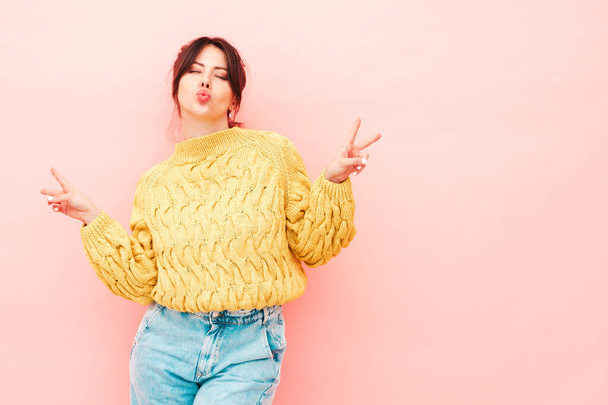 Young beautiful smiling female in trendy summer yellow hipster sweater and  jeans. Sexy carefree woman posing near pink wall in studio. Positive model having fun.Shows peace sign - Φωτογραφία, εικόνα