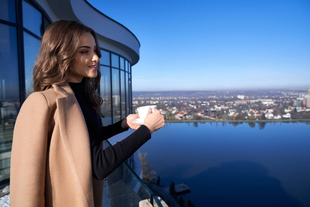 Gorgeous woman drinking coffee while standing on balcony - Фото, изображение