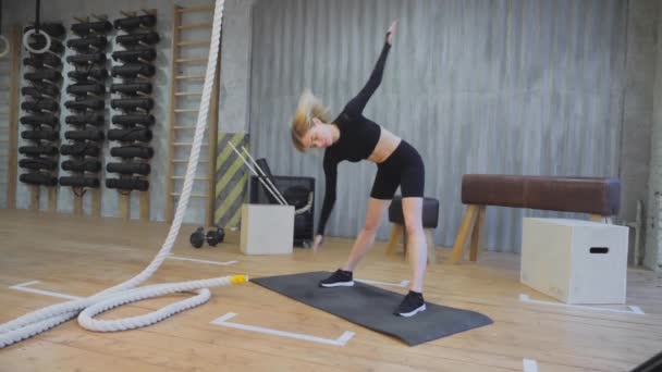 Sportieve blonde vrouw die sport doet warming-up voor de training, gekleed in sportieve zwarte kleren, lichte fitnessruimte - Video