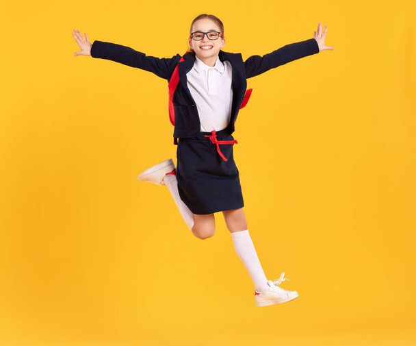 Full body of happy preteen schoolgirl in black uniform and glasses with backpack jumping high against yellow background - Photo, Image