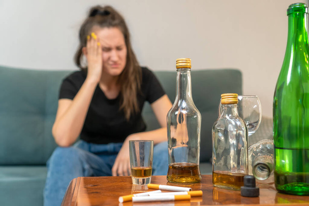 Betrunkene dunkelhaarige Frau sitzt auf dem Sofa und hält ihren Kopf, konzentriert auf leere Flaschen, Zigaretten. Gesellschaftliches Problem des Alkoholismus - Foto, Bild