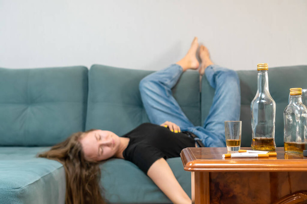 Humalainen nuori tummatukkainen nainen nukkuu sohvalla juhlien jälkeen, tyhjät pullot ovat pöydällä. Keskity pulloihin - Valokuva, kuva