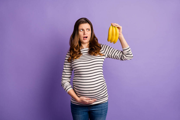 Photo portrait of pregnant upset woman holding looking at bananas isolated on vivid violet colored background - Фото, изображение