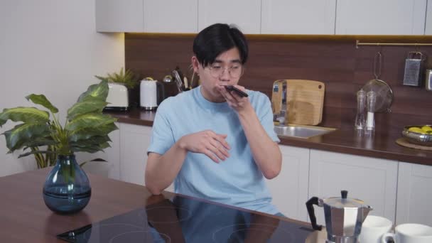 Mosolygó vietnámi férfi hangüzenetet vesz fel az otthoni konyhában. Boldog fiatal srác használja a telefont a közösségi hálózathoz és a hangvezérléshez. Közelkép, lassított felvétel 4k - Felvétel, videó