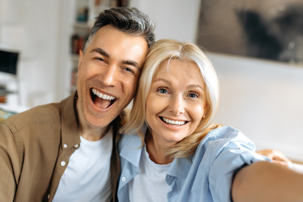 Portrait of beautiful cheerful marriage mature couple in casual clothes, Caucasian husband and wife taking selfie on smartphone, having fun together, looking at camera, smiling - Fotografie, Obrázek