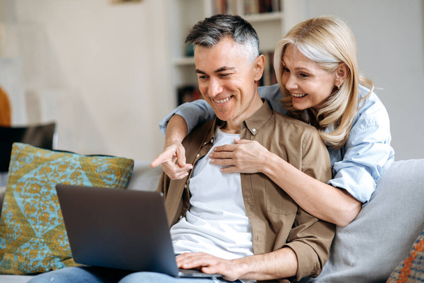 Joyful mature caucasian couple spend time at home, sitting on sofa in casual stylish wear, using laptop for online conference with friends or family, browsing internet, gesturing and smiling friendly - Foto, imagen