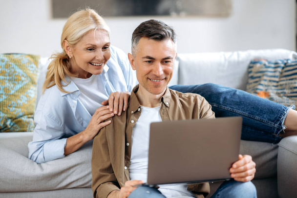 Happy caucasian family couple, mature wife and husband at home on a sofa, in casual wear, using laptop, browsing internet, social networks, chatting with friends or family, looking for household goods - Foto, immagini