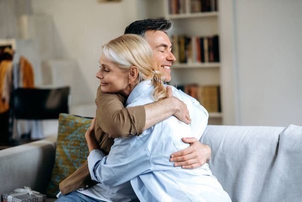 Счастливая любящая старшая кавказская семья, счастливые муж и жена, одетые в повседневную стильную одежду, сидящие дома на диване в гостиной, обнимающиеся, закрытые глаза, проводящие время вместе, улыбающиеся - Фото, изображение