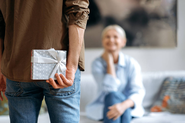 A present from husband on a special day. A loving husband in casual stylish clothes, holds a beautiful small box behind his back, prepares to give a surprise his beloved wife, a small anniversary gift - Photo, Image