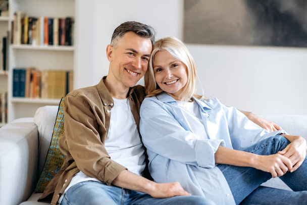 Portret przyjemnej szczęśliwej dojrzałej kaukaskiej pary małżeńskiej relaksującej się na kanapie w salonie w domu, spędzającej razem czas, ubranej w stylowe ubrania, uśmiechniętej do kamery, szczęśliwej razem - Zdjęcie, obraz