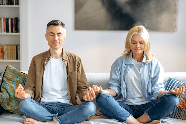 Reifes Liebespaar in lässiger Kleidung, das einen gesunden Lebensstil führt, zu Hause Yoga auf der Couch macht, im Wohnzimmer in Lotusposition sitzt, meditiert, die Augen geschlossen, lächelnd. Gelassenheit - Foto, Bild