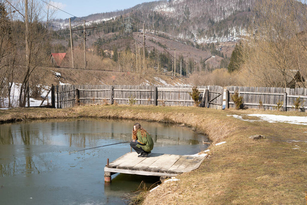 Семейная рыбалка Молодые люди сидят с удочками на деревянных пирсах у озера, покрытого тонким льдом. Рыбалка форели. Карпаты. Украина - Фото, изображение