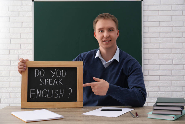 Teacher holding small chalkboard with inscription Do You Speak English? at table in classroom - Foto, imagen