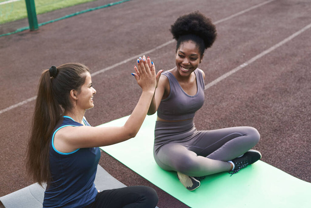 Two female athletes high-five while sitting on a yoga mat outdoors. Teamwork in sports training - Photo, Image