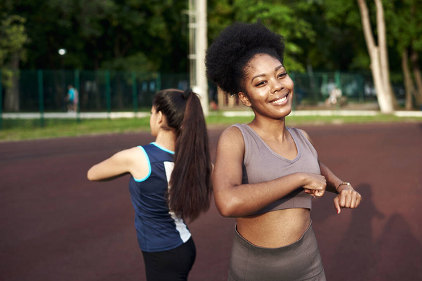 Két fitt, mosolygós fiatal nő együtt edz a parkban nyújtózkodik és nyújtja a kezét, hogy növelje a rugalmasságát. - Fotó, kép