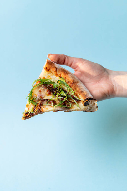 Woman is holding a slice of pizza with fresh arugula on blue studio background, close up, studio shot - Photo, Image