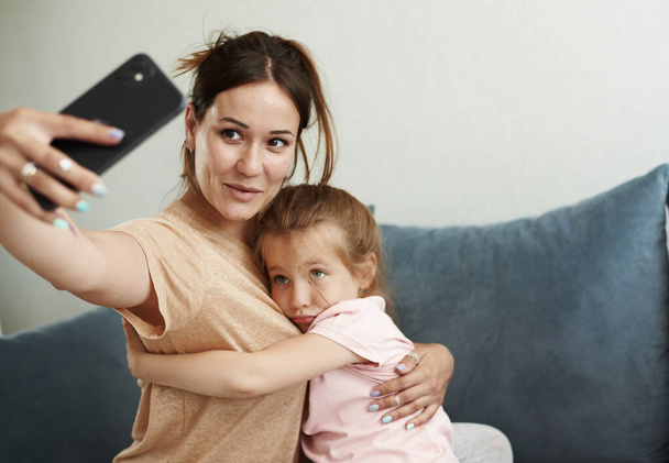 Joven madre caucásica y su pequeña hija linda tomar selfie en el teléfono, mostrando sus lenguas. Tiempo libre divertido en casa juntos - Foto, Imagen