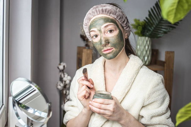 Young beautiful woman in a dressing bathrobe is applying a green cosmetic mask to her face. Concept of skin care and lifestyle. - 写真・画像
