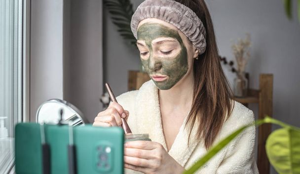 Pretty female blogger is putting a cosmetic green mask on her face in front of a mobile phone camera. - 写真・画像