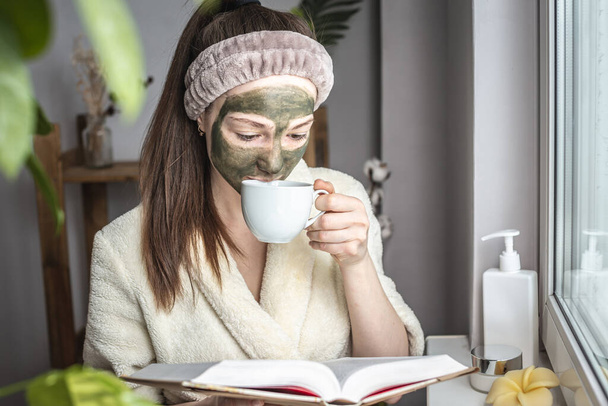 Pretty young woman in a bathrobe and with a green cosmetic mask on her face is reading a book and drinking tea. Concept of relaxation, beauty treatments, skin care and lifestyle - 写真・画像