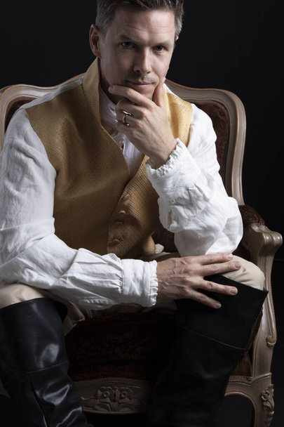 Komea Regency mies yllään kulta liivi, ja housut ja istuu punainen sametti tuoli pimeässä huoneessa. - Valokuva, kuva