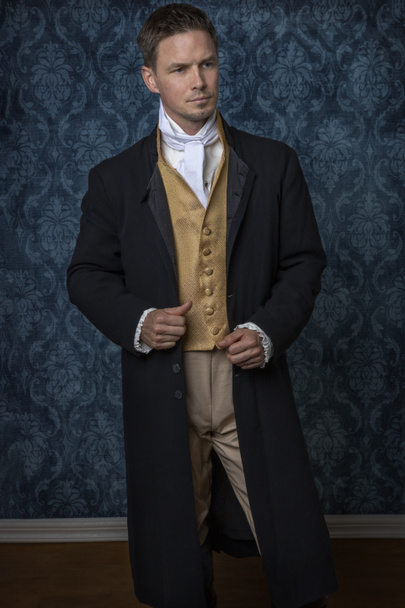 Un hombre guapo de Regency con un chaleco de oro, calzones, y una chaqueta negra y de pie en una habitación con papel pintado azul y un piso de madera.  - Foto, imagen