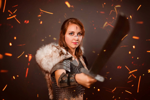 Medieval woman warrior in chain mail armor and polar fox fur on her shoulders standing in a fighting stance with a sword in her hands on a dark background, bokeh of blurry sparks in the foreground. - 写真・画像