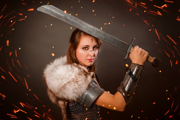 Medieval woman warrior in chain mail armor and polar fox fur on her shoulders standing in a fighting stance with a sword in her hands on a dark background, bokeh of blurry sparks in the foreground. - Photo, Image