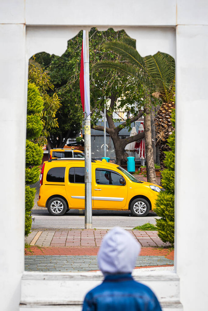 Концепция путешествия. Маленький мальчик смотрит через белую дверь, окно на желтое такси, движущееся через улицу к зеленым деревьям, пальмы в теплой жаркой стране - Фото, изображение