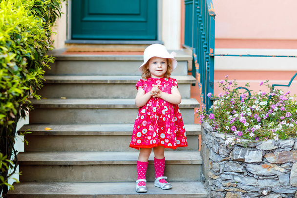 Portrait of beautiful little gorgeus lovely toddler girl in pink summer look clothes, fashion dress, knee socks and hat. Happy healthy baby child posing infront of colorful house. - Photo, Image