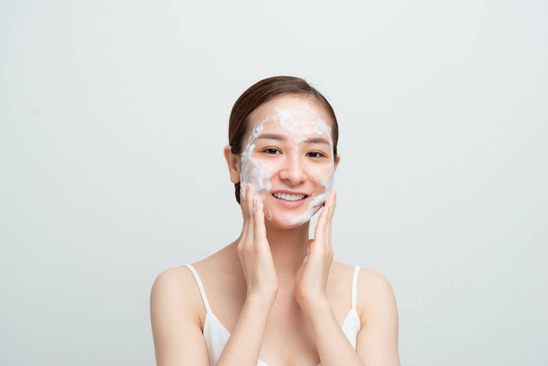Primo piano di felice donna asiatica sorridente utilizzando la schiuma detergente per la pelle quotidiana e guardando felice, sfondo bianco - Foto, immagini