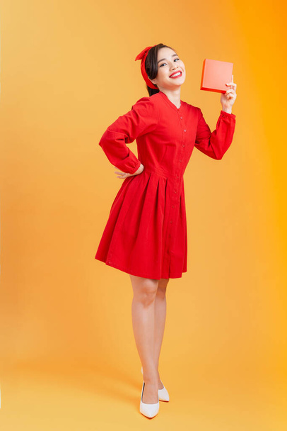 люди, рождество, день рождения и праздники концепция - счастливая молодая женщина в красном платье показ / проведение с подарочной коробкой на оранжевом фоне. - Фото, изображение