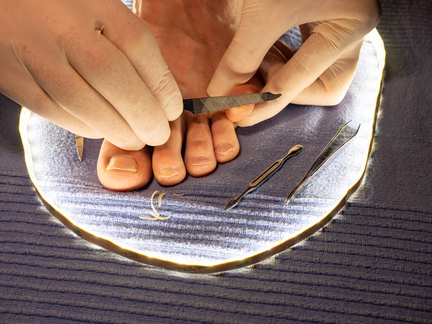 Las manos en guantes estériles blancos lijan las uñas de los pies con una larga lima puntiaguda en los dedos masculinos. Pedicura sobre toalla azul - Foto, Imagen