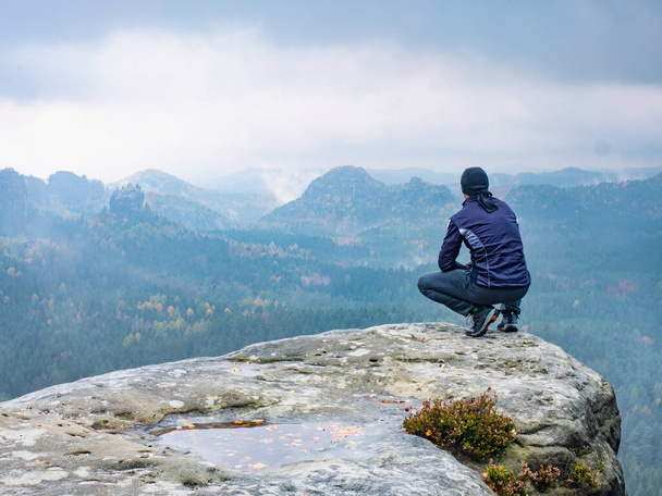 Un uomo seduto sulla cima di una montagna in una mattina nebbiosa. Sedetevi accovacciati sul bordo della vetta e godetevi le montagne vista valle durante la nebbia pesante - Foto, immagini