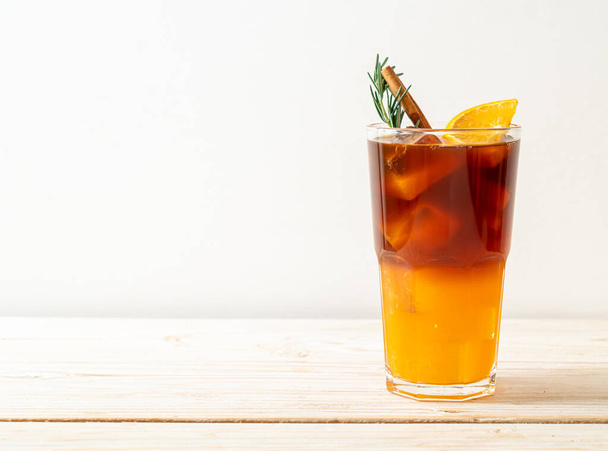 A glass of iced americano black coffee and layer of orange and lemon juice decorated with rosemary and cinnamon on wood background - Fotografie, Obrázek