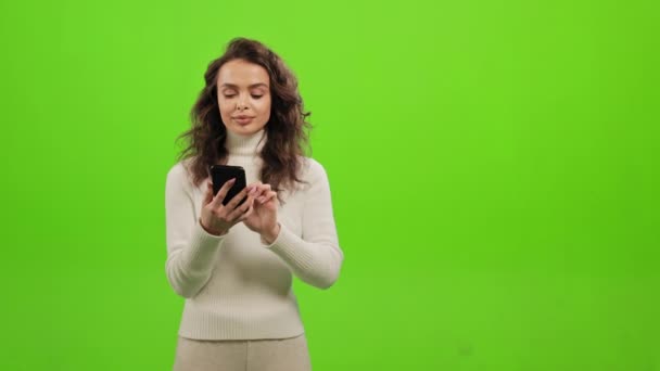 The woman is looking through social networks on her smartphone. She is raising her head and looking at the camera. She has an idea and raises her finger up. She is standing on a green background - 映像、動画