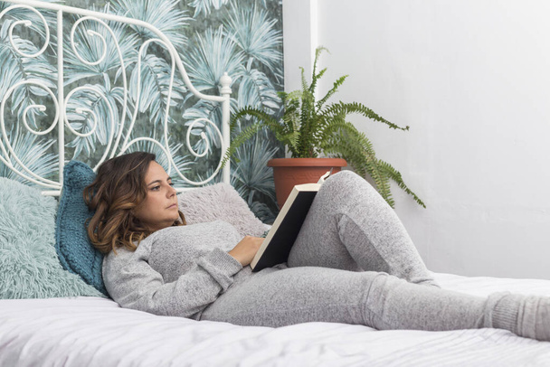 woman lying in bed reading a book, brunette woman in gray pajamas reads book lying on  bed with a white coverlet, next to the bed there is a fern - Foto, imagen