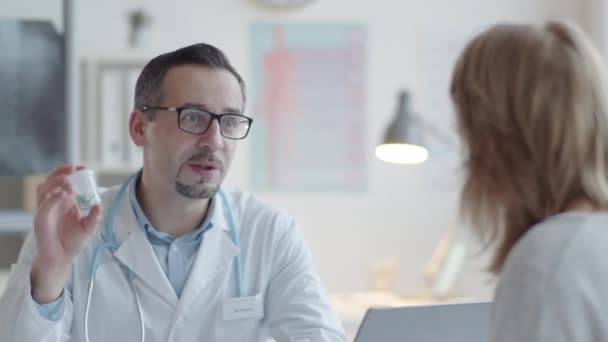 Мужчина-врач в лабораторном халате со стетоскопом на шее, дающий лекарство пациентке, объясняющий, как его принимать и выписывающий рецепт во время консультации в медицинском кабинете - Кадры, видео