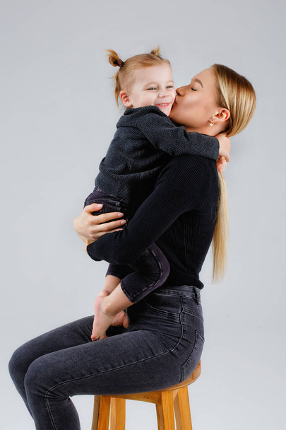 A mother embraces her daughter on a gray background in the studio with love. A woman and a child are blonde in dark clothes isolated in the background. - Photo, image