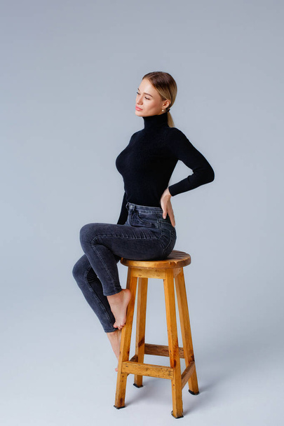 Portrait of a young blonde woman in black clothes sitting on a stool in the studio. Gray background. - Foto, Imagen