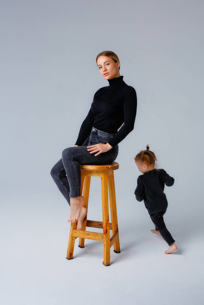 Porträt einer jungen Blondine in schwarzer Kleidung, die auf einem Hocker im Atelier sitzt. Ein kleines Mädchen rennt herum. Grauer Hintergrund. - Foto, Bild