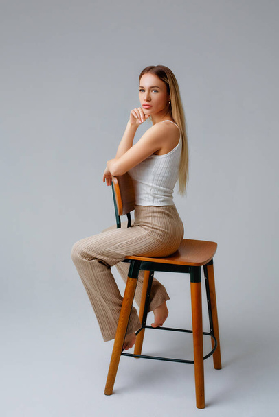 Uma jovem bela loira em uma camiseta branca elegante e calças de malha bege senta-se em uma cadeira em meia volta em um fundo branco no estúdio. - Foto, Imagem