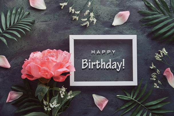 Happy birthday card with peony and palm leaves. Flowers, petals and exotic leaves around white frame on black, dark grey distressed textured background. Creative flat lay with natural floral design. - Photo, image