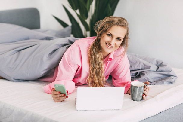 Una hermosa chica está acostada en la cama con una camisa rosa, sosteniendo una taza de café en la mano y usando una computadora portátil para ver contenido en línea, capacitación en línea, trabajo en línea - Foto, Imagen
