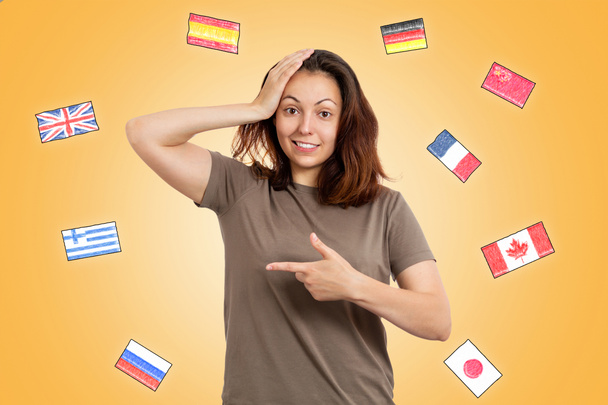 Die junge lächelnde, nachdenkliche Studentin wählt eine Sprache zum Lernen, zeigt mit dem Finger nach links. Gelber Hintergrund mit Flaggen verschiedener Länder. Das Konzept des Erlernens von Fremdsprachen. - Foto, Bild