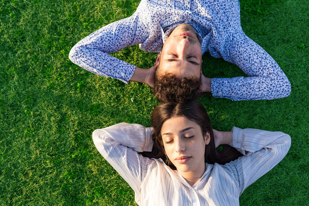 Top view of carefree young heterosexual couple resting sleeping on a beautiful green lawn with hands under heads. Two people relaxing enjoying together living the nature to healthy and better life - Photo, Image