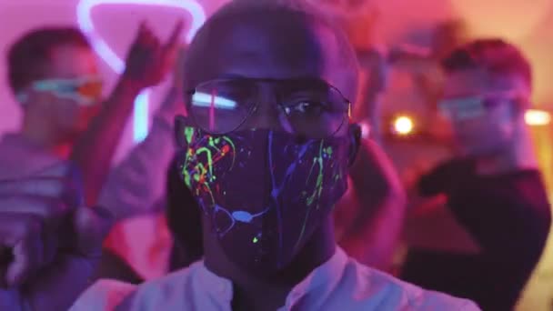 Nahaufnahme eines jungen schwarzen Mannes in Brille und Gesichtsmaske mit Neon-Farbspritzern, der auf einer Hausparty vor der Kamera tanzt - Filmmaterial, Video