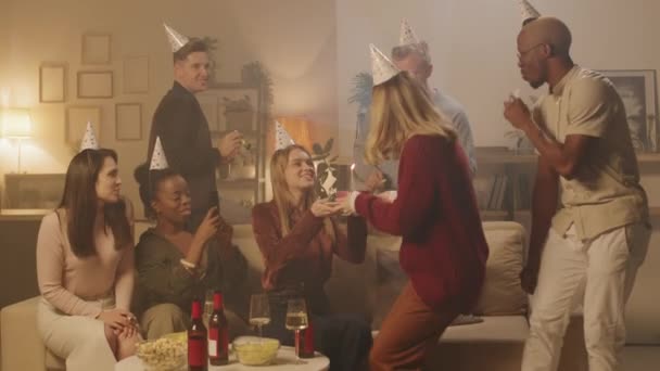 Střední záběr šťastné skupiny přátel foukání party rohy a tleskání, zatímco mladá žena foukání svíčku na narozeninový dort - Záběry, video