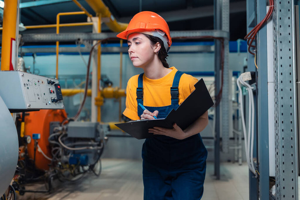 Industrial production concept. A young caucasian woman in uniform and helmet with a folder and a pen in her hand, writing meter readings. In the background-boiler equipment and pipes. - Photo, Image